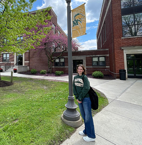 girl in green marywood sweatshirt outside leaning against pole that has gold marywood banner on it